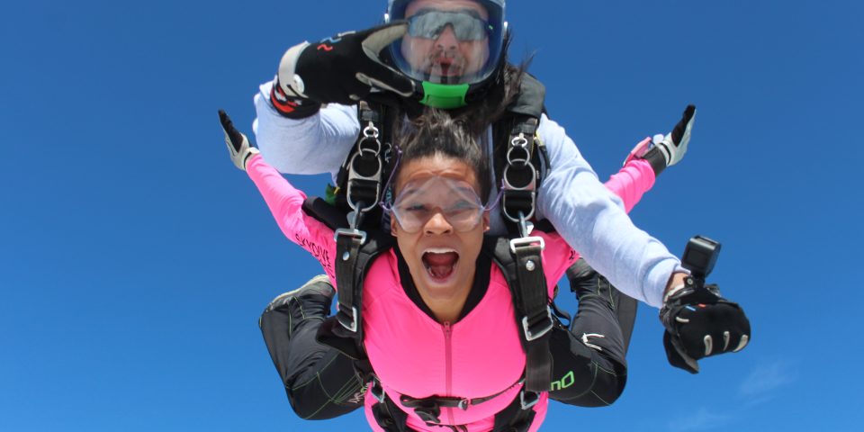A girl in a pink jumpsuit smiles during free fall on a tandem skydive.